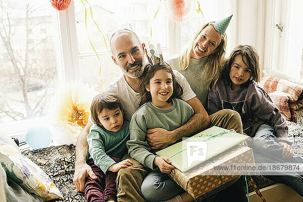 Porträt eines glücklichen Geburtstagskindes  das mit Geschenken und Familie auf einem Nischensitz zu Hause sitzt