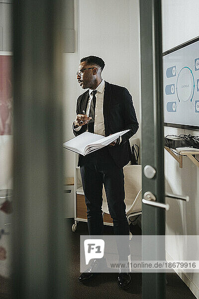 Mature male entrepreneur holding notebook during business meeting at office seen through doorway