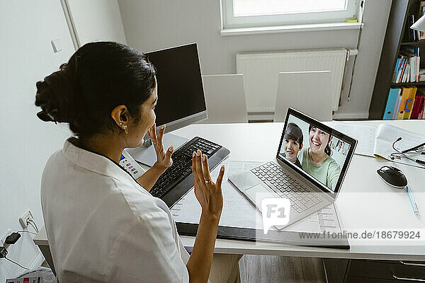 High angle view of female pediatrician waving patients over video call through laptop at desk in clinic