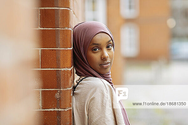 Portrait of young woman in hijab leaning against brick wall