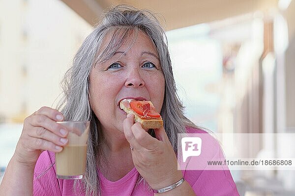 Nahaufnahme einer älteren weißhaarigen Frau  die Kaffee mit Milch trinkt und einen Toast mit Schinken und Tomaten isst