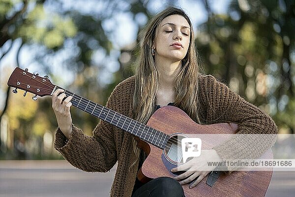 Verträumte und berührte zarte blonde Frau  schließt die Augen und spielt Gitarre mit zufriedenem freundlichem Lächeln in einem Park