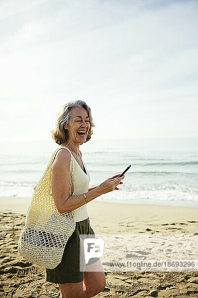 Happy woman with mesh bag holding smart phone at beach