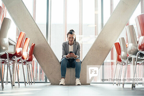 Lächelnde Geschäftsfrau sitzt mit Smartphone auf einer Säule im Büro