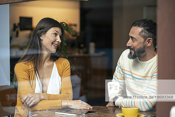 Couple talking to each other sitting in cafe seen through glass window