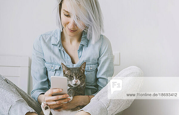 Smiling woman holding cat and using smart phone at home