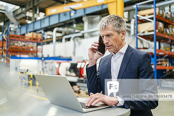 Businessman talking on smart phone using laptop at factory