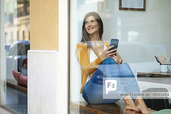 Smiling woman with mobile phone sitting in cafe seen through glass window