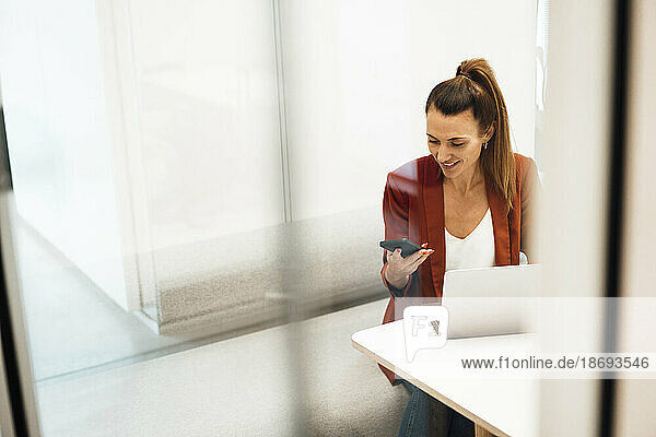 Businesswoman with laptop using smart phone sitting at desk in office