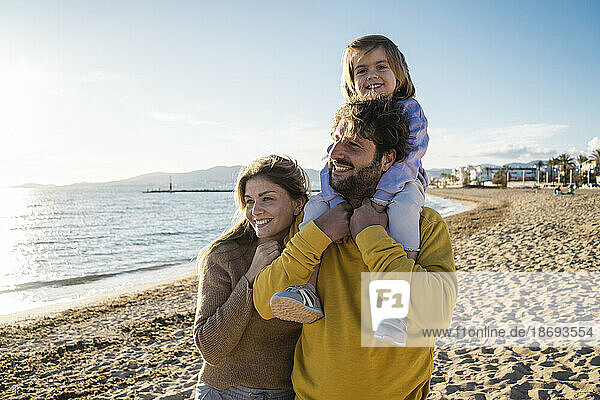 Smiling daughter sitting on father's shoulders by woman at beach