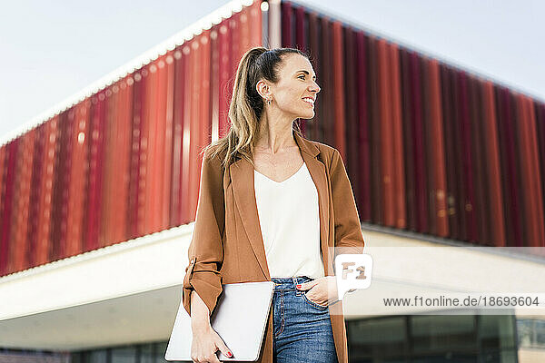 Lächelnde Geschäftsfrau mit der Hand in der Tasche  die vor einem modernen Gebäude steht