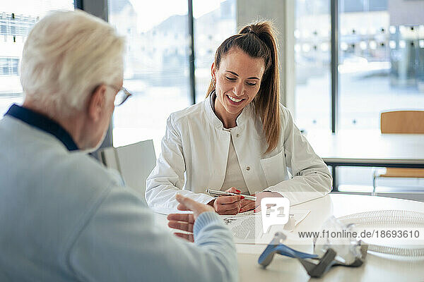 Smiling doctor advising senior patient sitting at desk in clinic