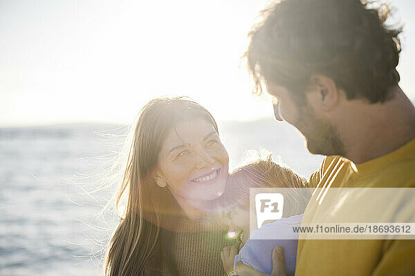 Smiling woman looking at man holding daughter at beach on sunny day