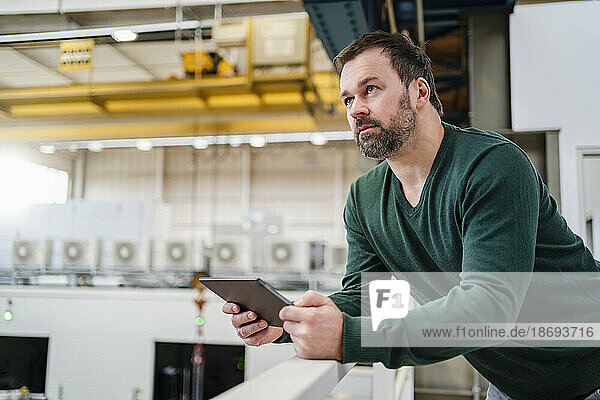 Mann hält Tablet-PC in der Hand und lehnt an Geländer in Fabrik