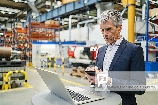 Businessman using smart phone by laptop on table at factory