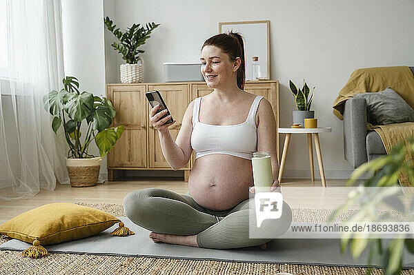 Lächelnde schwangere Frau  die zu Hause auf einer Trainingsmatte sitzt und ihr Smartphone benutzt