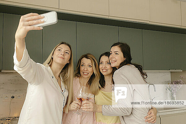 Woman taking selfie through smart phone with friends in kitchen at home