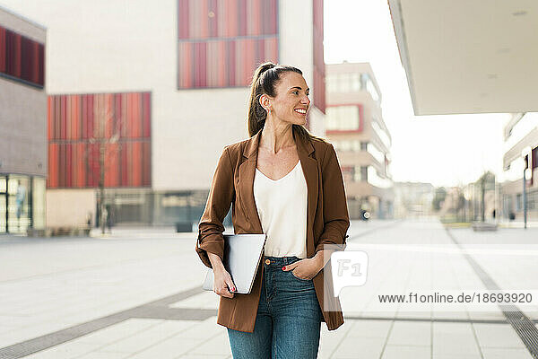 Smiling businesswoman holding laptop and walking on footpath