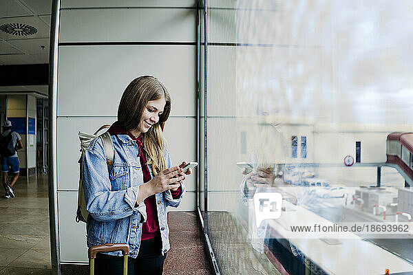 Young woman with suitcase using phone by glass window at airport