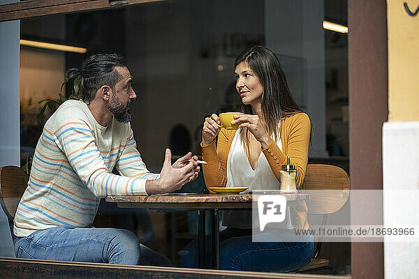 Man talking to woman holding cup sitting in cafe seen through glass window
