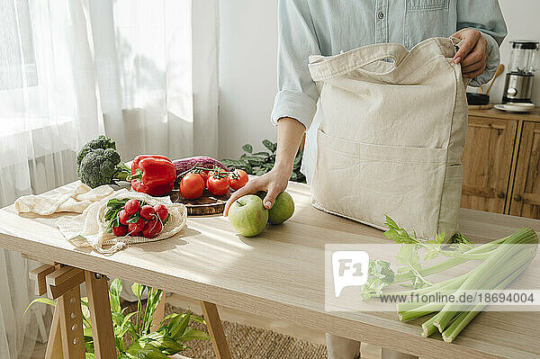 Woman keeping apples by reusable bag on table at home