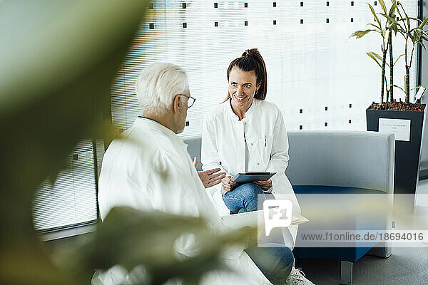 Smiling doctor discussing with senior colleague sitting on couch at hospital
