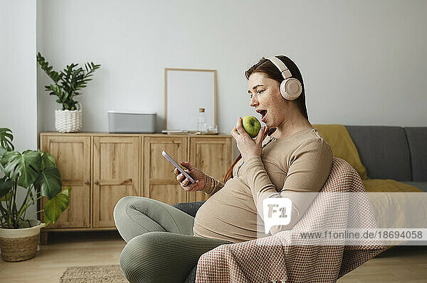 Schwangere Frau isst Apfel und benutzt Smartphone  während sie zu Hause auf einem Stuhl sitzt