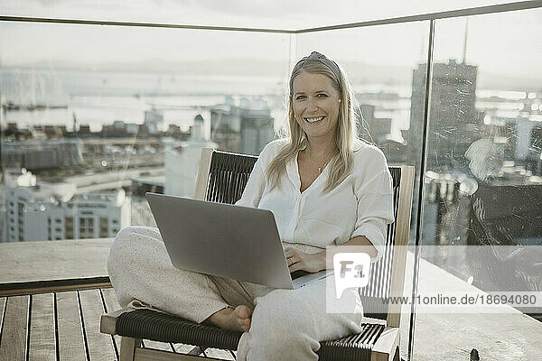 Happy young freelancer with laptop sitting on chair on rooftop
