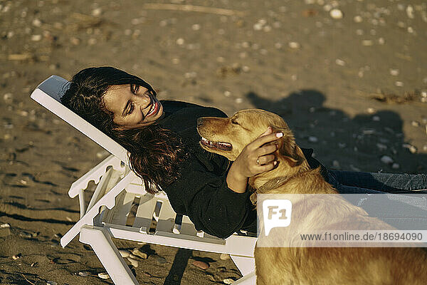 Smiling woman lying on deck chair and stroking dog at beach