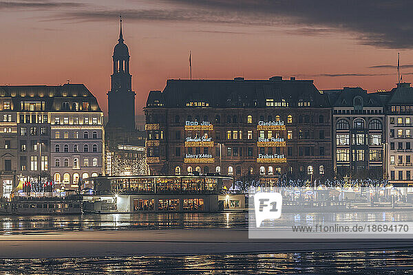 Germany  Hamburg  Christmas decorations hanging on city buildings at dusk with Alster Lake in foreground