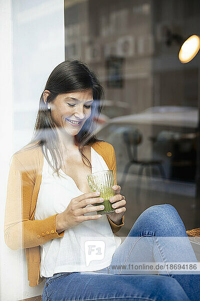 Lächelnde Frau hält gesundes Getränk in der Hand und sitzt im Café  gesehen durch die Glasscheibe