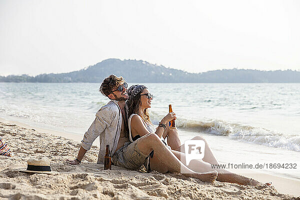 Happy couple enjoying drinks sitting on sand at beach