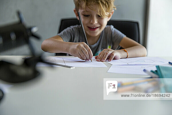 Lächelnder Junge  der am Schreibtisch mit Bleistift auf Papier zeichnet