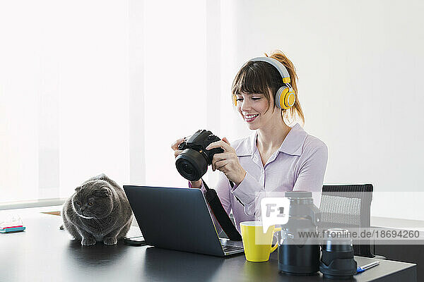 Happy businesswoman wearing wireless headphones sitting with camera at desk