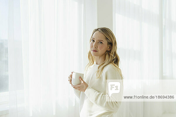 Blond woman holding coffee cup by translucent curtain
