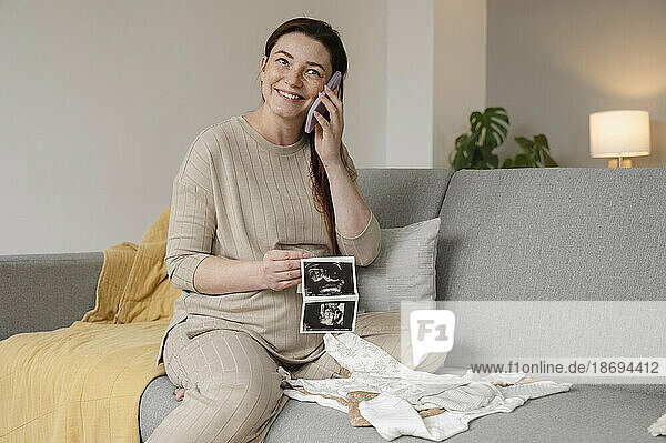 Happy pregnant woman talking on smart phone sitting with ultrasound images at home