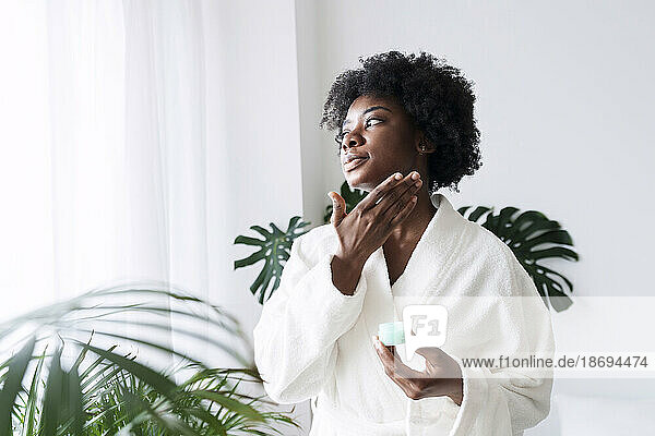 Frau im Bademantel trägt zu Hause Feuchtigkeitscreme auf das Gesicht auf