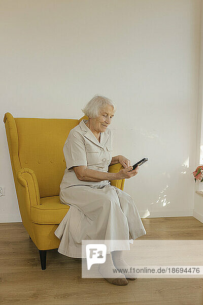 Elderly woman using smart phone sitting on armchair at home