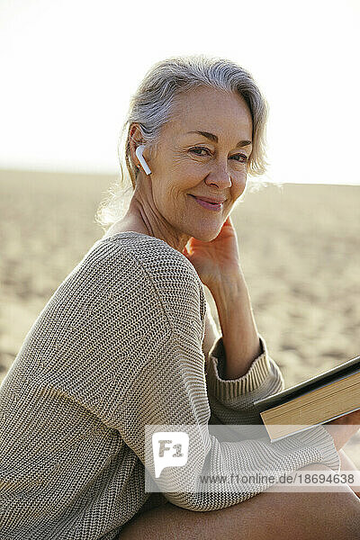 Smiling mature woman wearing wireless in-ear headphones at beach