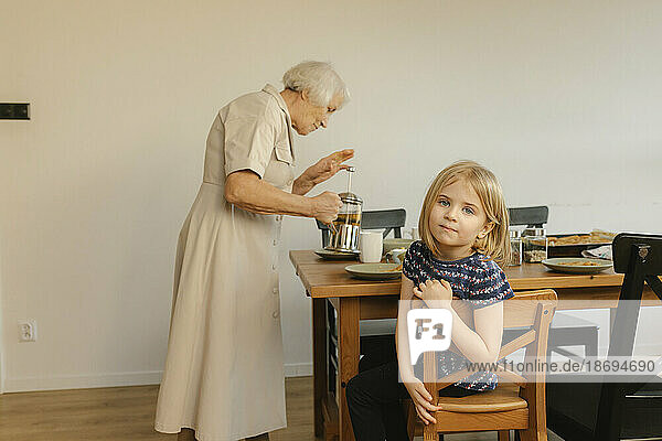 Girl sitting on chair with grandmother preparing tea in background at home