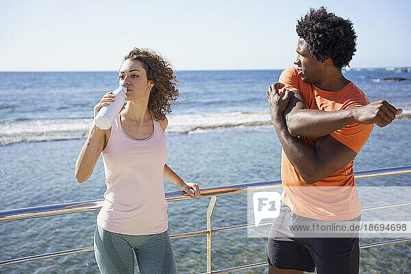 Man exercising with girlfriend drinking water by railing in coastal area