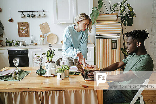 Woman talking to man working with laptop on dining table at home