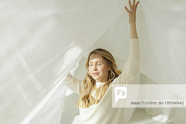 Blond woman with eyes closed and arms raised under white translucent curtain
