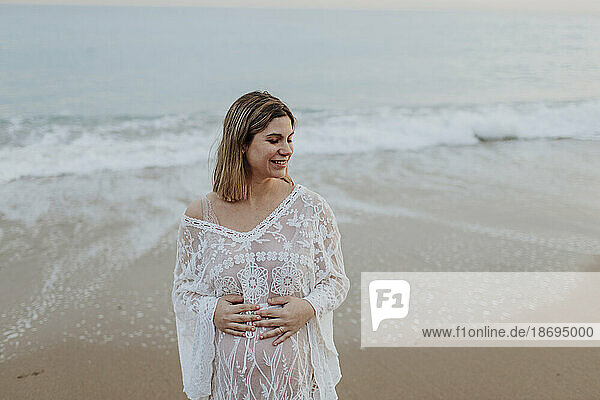 Lächelnde schwangere Frau mit den Händen auf dem Bauch steht am Strand