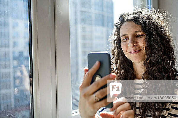 Smiling woman using smart phone near window
