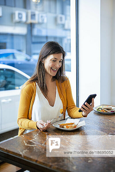 Happy woman using smart phone sitting with food on table at cafe