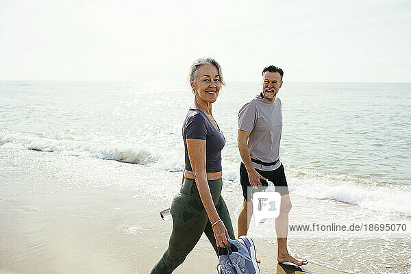 Smiling woman walking with man holding sports shoes at beach