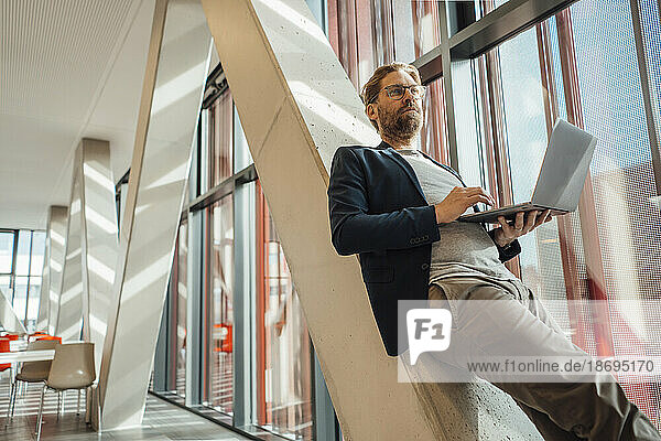 Businessman holding laptop leaning on column in building
