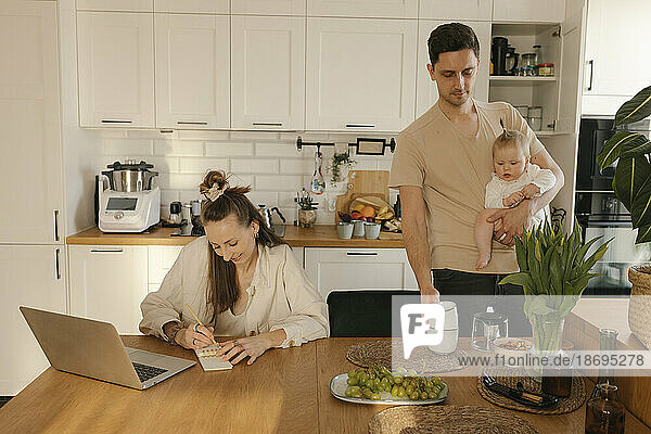 Woman working on laptop by man carrying daughter doing chores at home