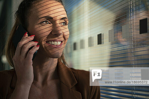 Glückliche Geschäftsfrau  die im Büro an der Wand mit dem Smartphone spricht